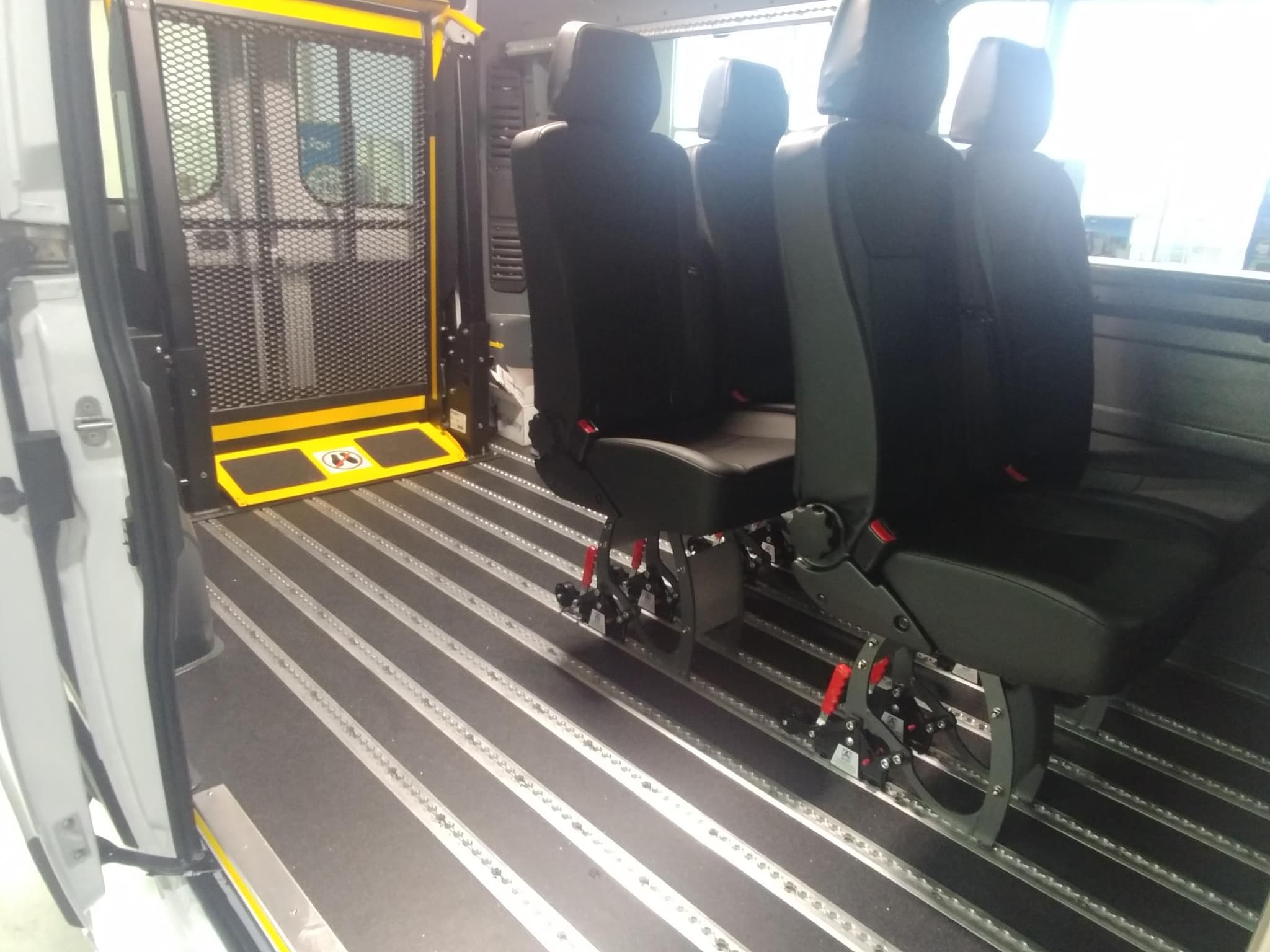 Inside a ProMaster conversion looking toward the side door: the powered wheelchair lift folded upright in the doorway, and four black removable passenger seats mounted on aluminium floor tracks with red quick-release handles