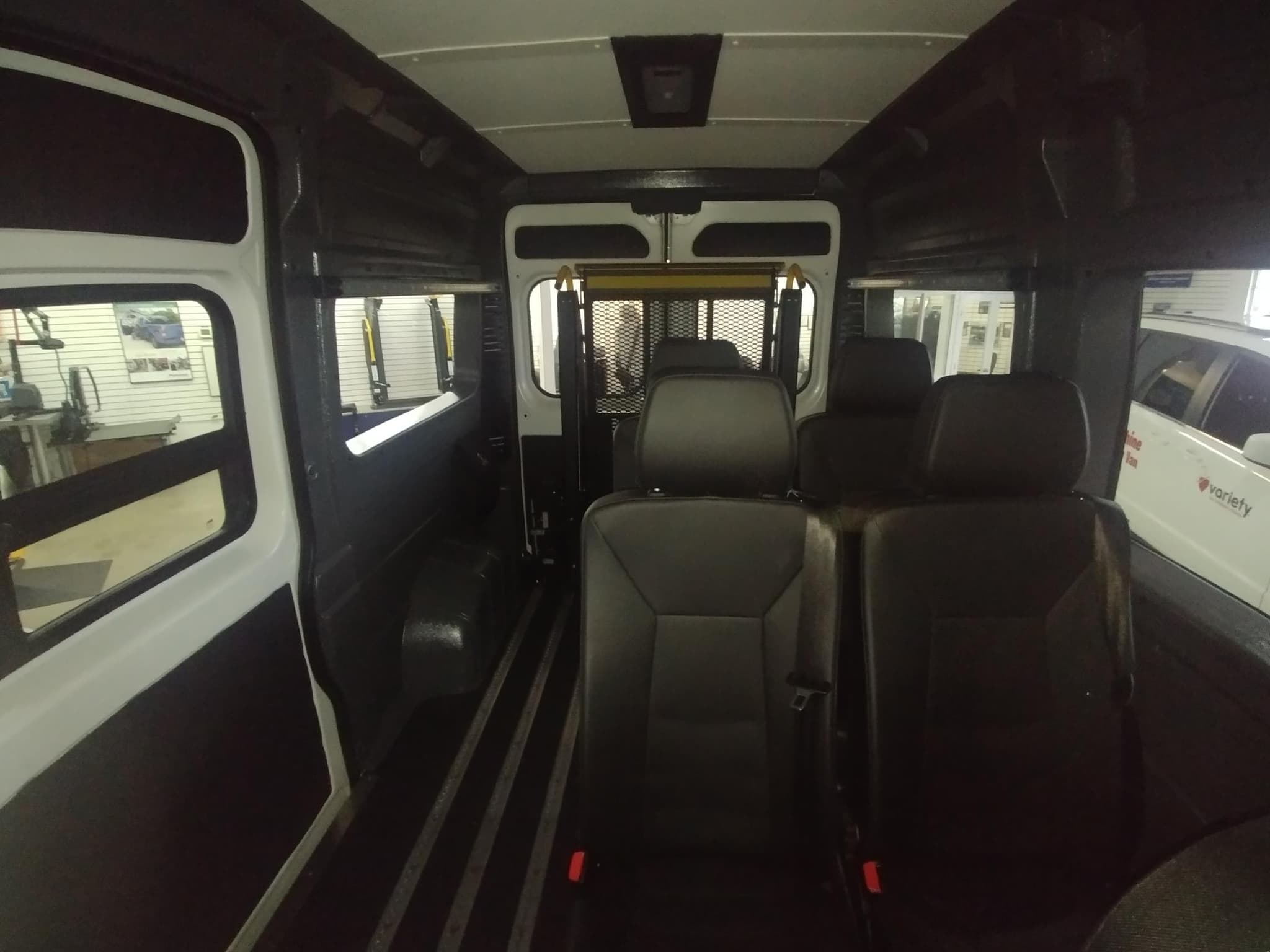 Looking down the high roof ProMaster cabin from the front, black seats on either side of a clear centre aisle, with the wheelchair lift and its mesh safety gate at the rear doors