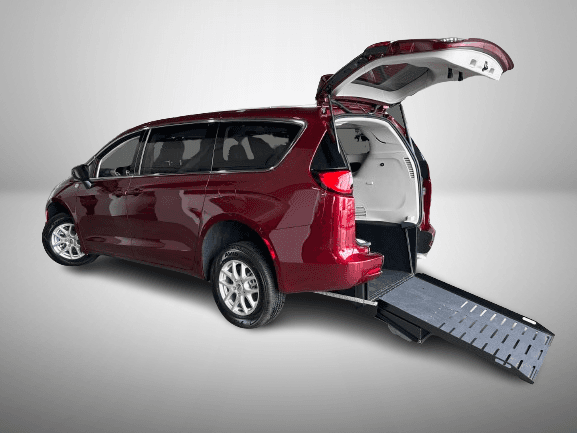 The same red Grand Caravan from the rear driver's side with the liftgate up and the ramp folded out to the ground, photographed against a plain studio background
