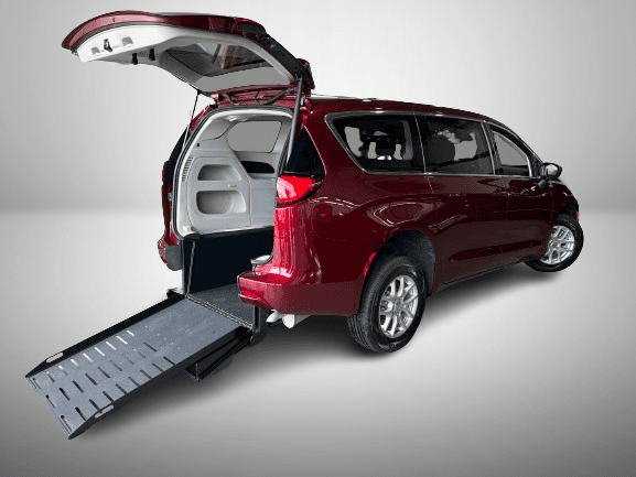 Red Chrysler Grand Caravan seen from the rear passenger side with the liftgate raised and the manual foldout ramp extended to the ground, photographed against a plain studio background