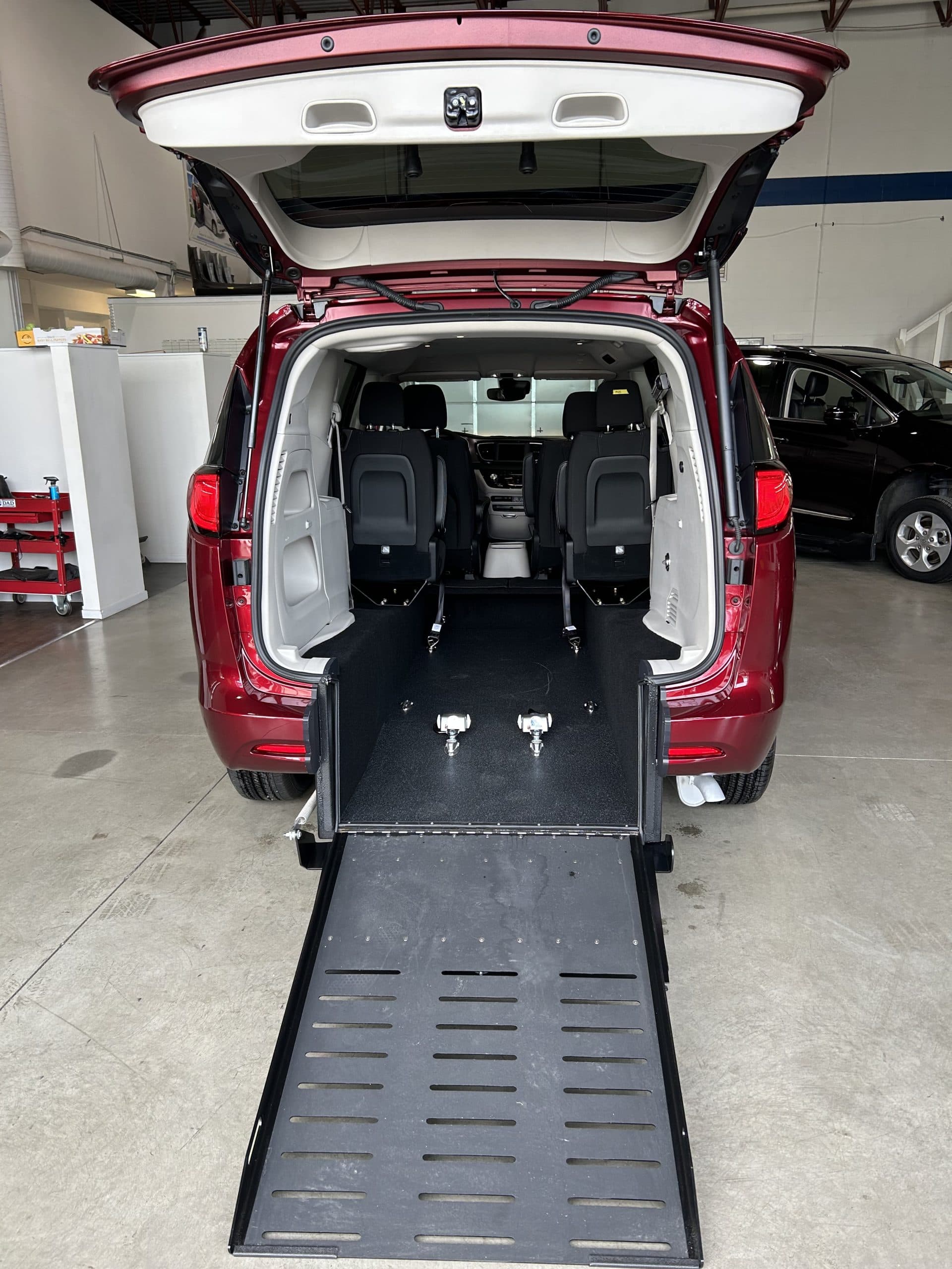 Red Chrysler Grand Caravan photographed in the Sidewinder shop, rear liftgate raised and the manual foldout ramp lowered to the floor, showing the flat black non-slip lowered floor and the second row bucket seats set outboard