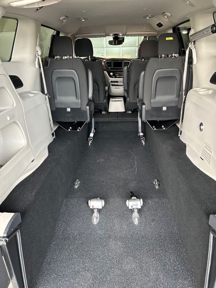 Inside a Sidewinder RAV rear-entry conversion looking forward from the open liftgate: black non-slip flooring running the length of the lowered floor, two chrome wheelchair securement anchors set into the floor, and black bucket seats against light grey trim