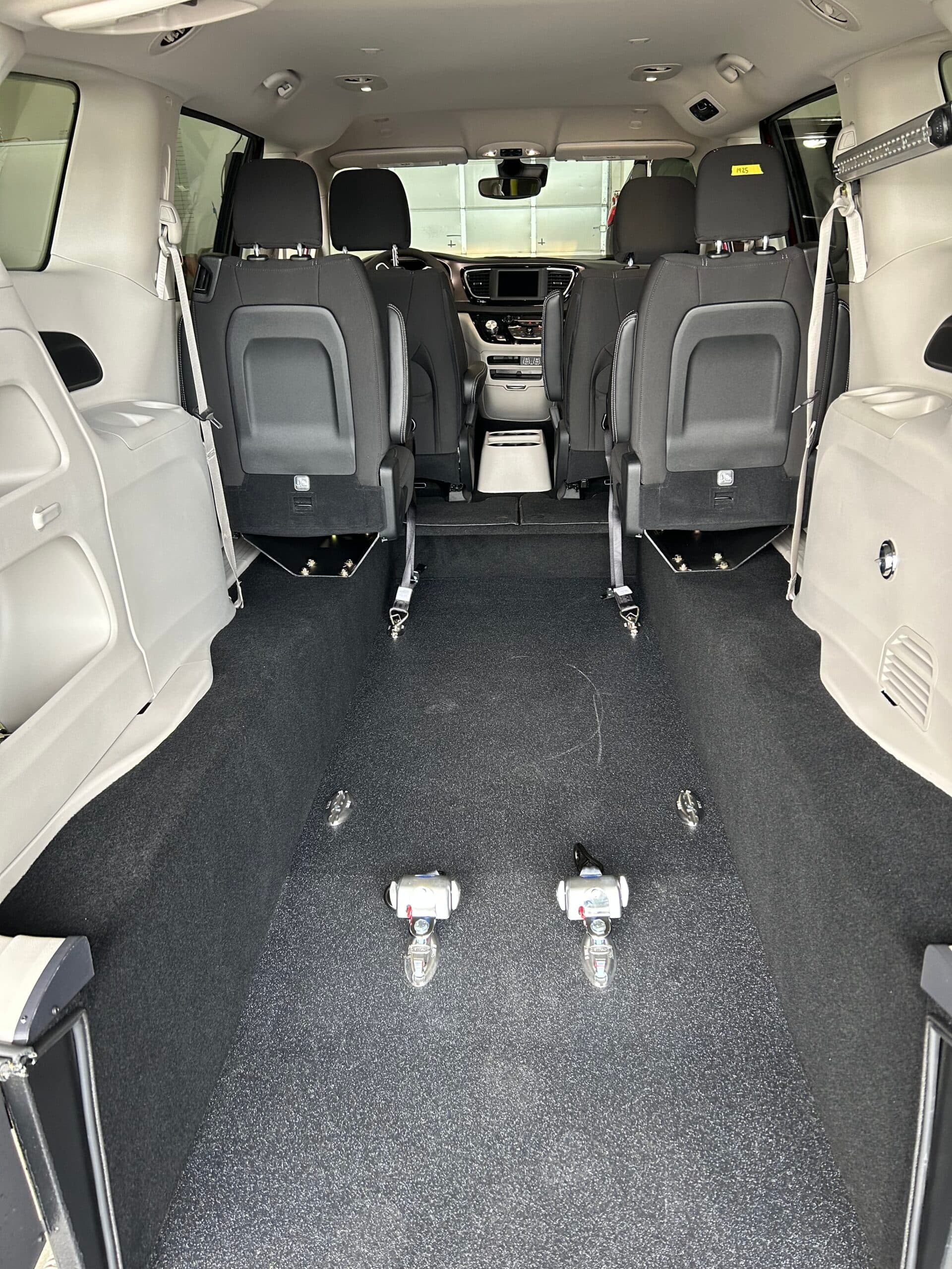 Inside a Sidewinder RAV rear-entry conversion looking forward from the open liftgate: black non-slip flooring running the length of the lowered floor, four wheelchair securement anchors set into the floor, and black bucket seats against light grey trim
