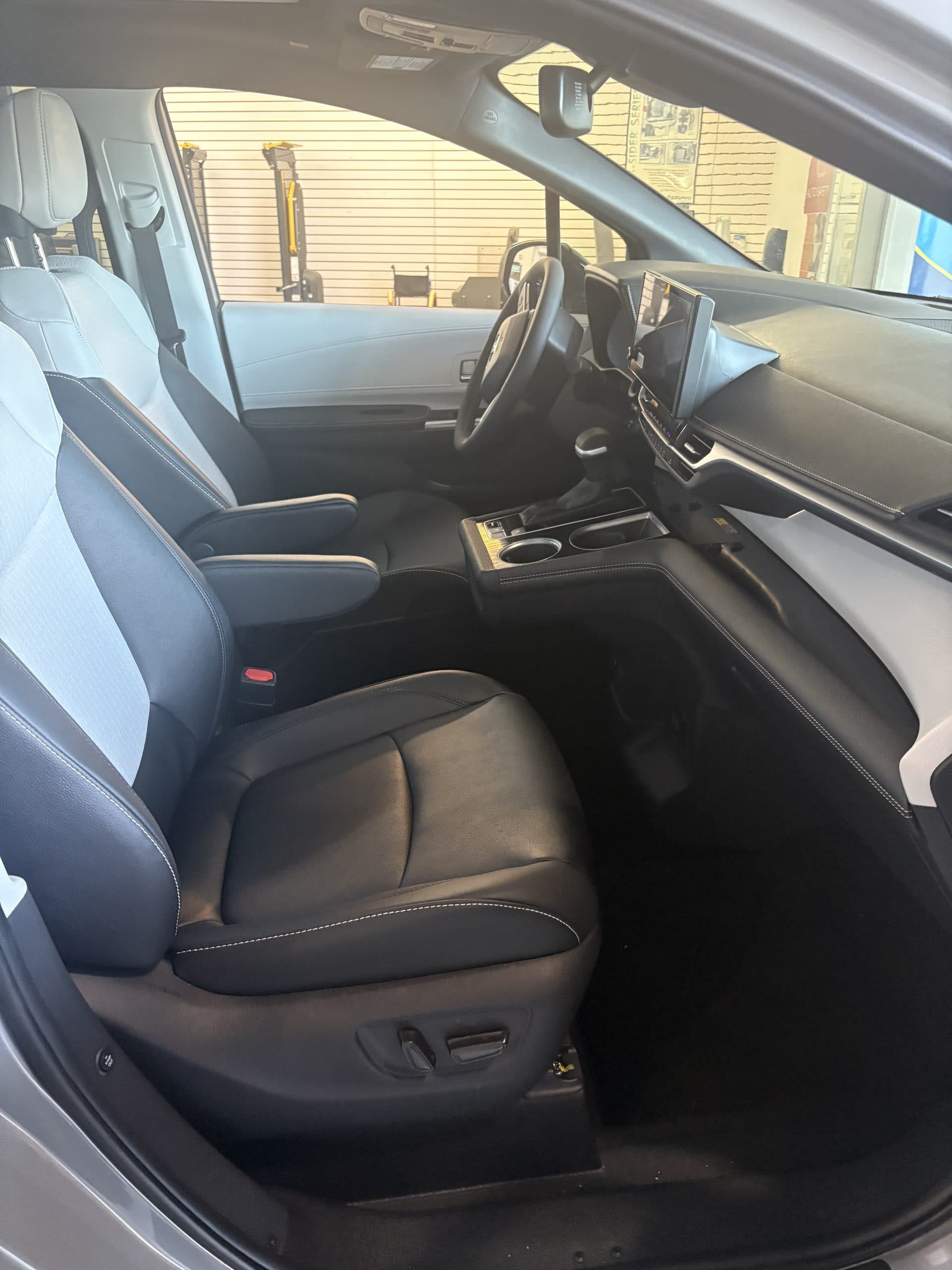 Front passenger side of the Sienna from the open door, showing the black leather seat, the centre console and the dashboard, with a wheelchair visible in the workshop outside
